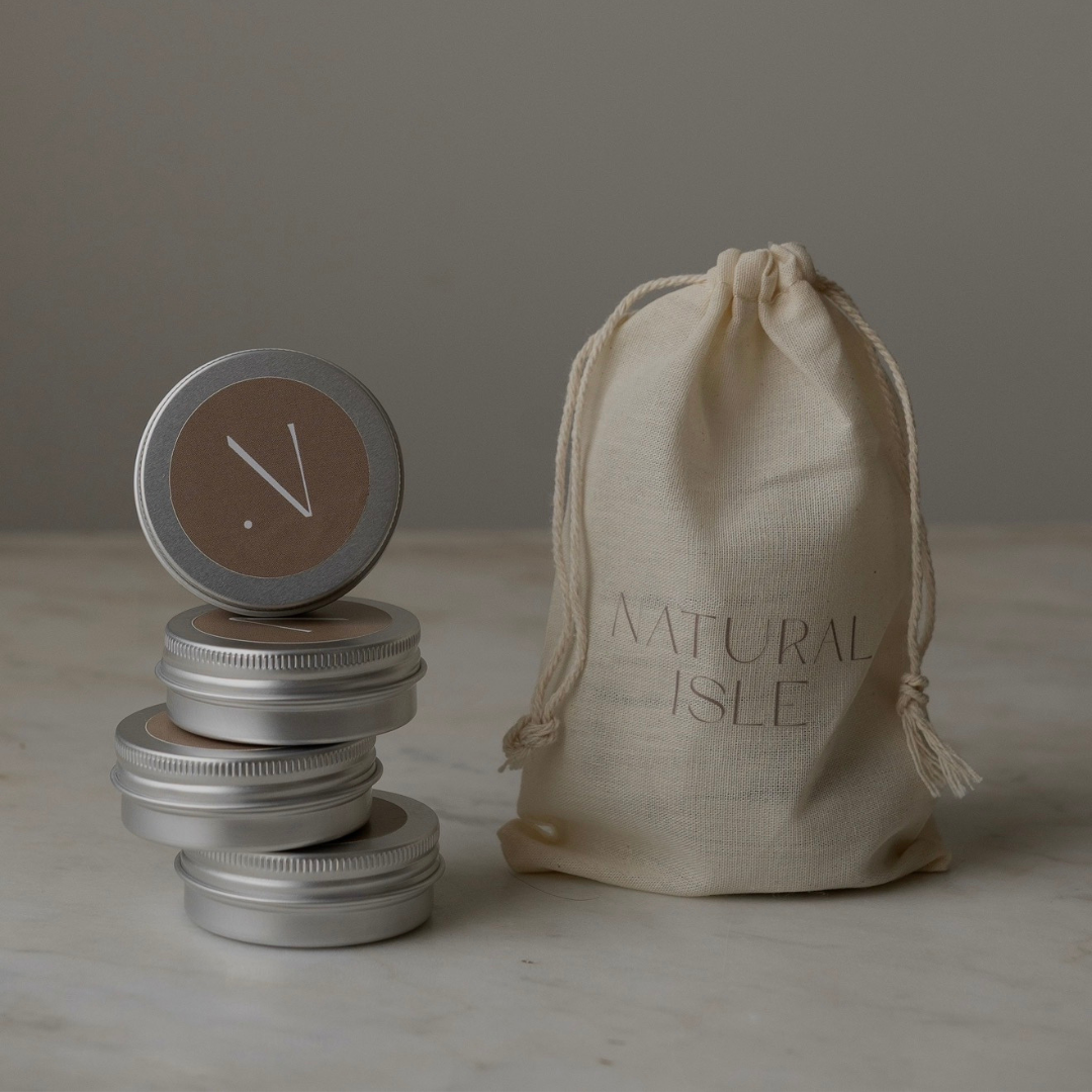 Candle tins with decorative lids on a marble surface, featuring a 'Natural Isle' drawstring bag.