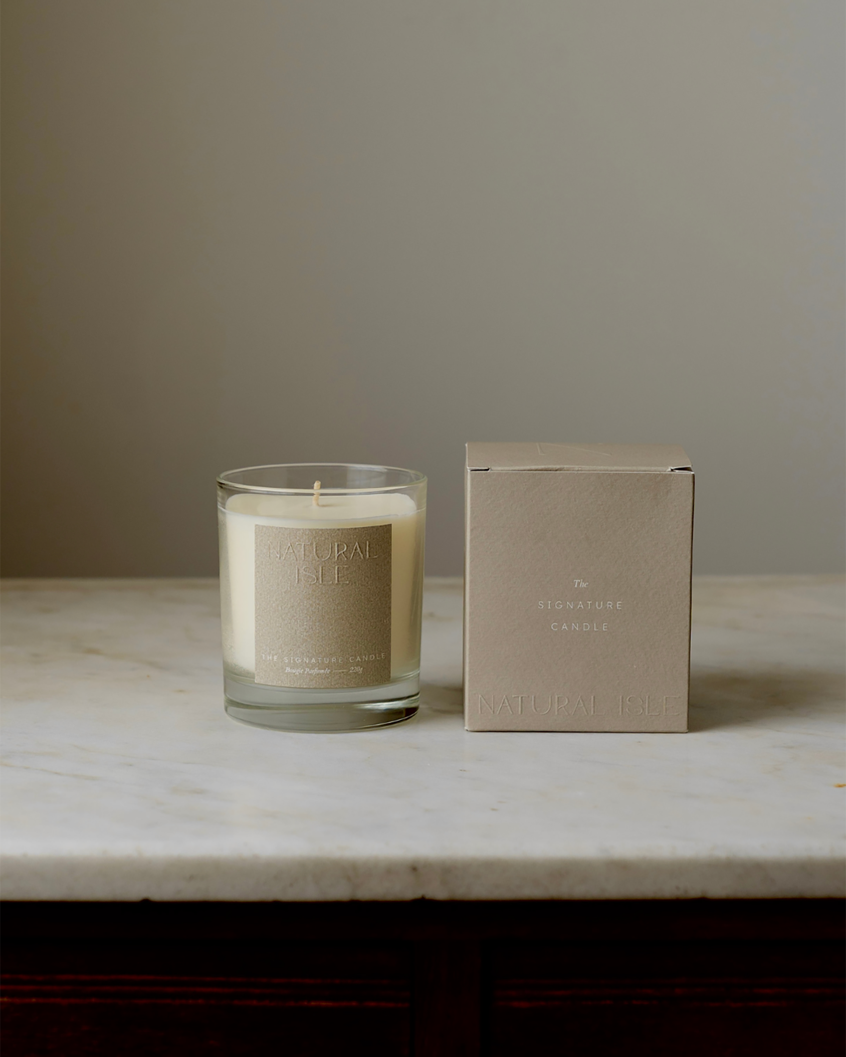 Natural Isle Signature Candle, Glass candle with a beige label on a marble surface next to its packaging box.