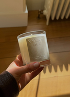 Natural Isle Signature Candle, Hand holding a candle labeled 'Nature Isle' with wooden floor and delicate natural sunlight in the background